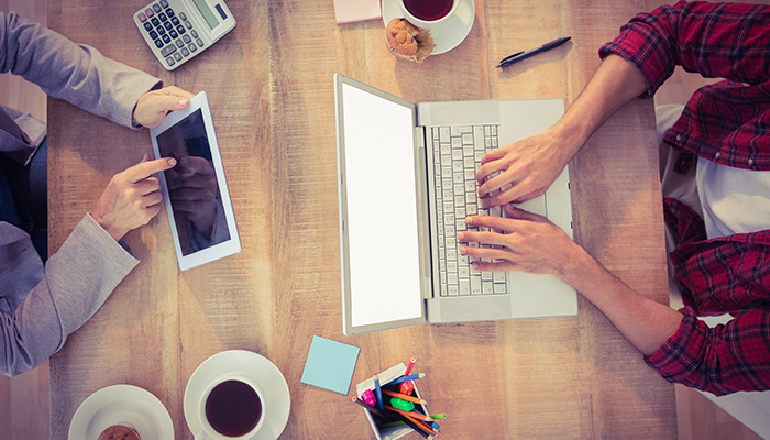 People sitting in a coffee house with two cups of coffee, a calculator, pens, paper, tablet, and a laptop on the table